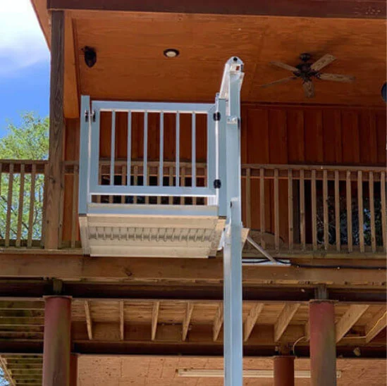 Shuttle + Cargo Lift Outdoor home elevator on a wooden house, connecting upper balcony to the ground. White metal framework and wooden ceiling fan visible.