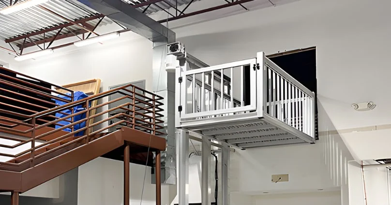Modern home elevator with safety rails accessing an upper floor in an industrial-style space, near a staircase with brown metal railing.
