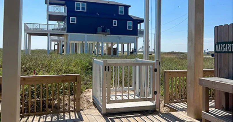 Elevated beach house with stilts and spacious decks, home elevator in foreground for easy access, surrounded by lush grass and blue sky.